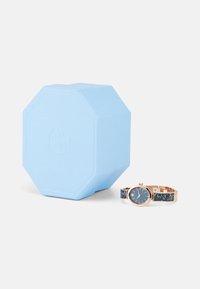 Reloj de pulsera de oro rosa con esfera azul y pulsera azul brillante, al lado de una caja de cartón hexagonal de color azul claro.