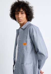 Striped work jacket in blue and white, featuring a chest pocket with an orange tag, button closure, and a relaxed fit. Soft cotton fabric.