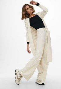Cremefarbene, gerippte Strickjacke über einem schwarzen Langarmoberteil, kombiniert mit hellbeigen Hosen. Klobige beige und schwarze Sneakers.