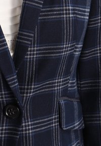 Blazer bleu marine à motif à carreaux blancs et bleu clair, bouton à l'avant et petite poche à rabat. Tissu blanc visible en dessous.