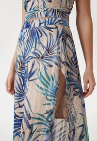 Una falda fluida hasta la rodilla con una base beige y patrones de hojas tropicales azules y verdes, de tela transparente y con una abertura alta en el muslo.