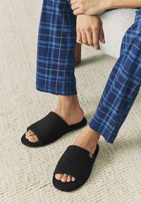 Chaussons noirs texturés avec de larges lanières ouvertes ; portés avec un pantalon de pyjama à carreaux bleus. Les chaussons présentent une semelle plate et un design minimaliste.
