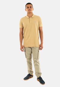 Camisa polo de algodón beige con cuello, combinada con pantalones cargo beige claros y zapatillas navy con detalles en naranja, de pie frente a un fondo blanco.