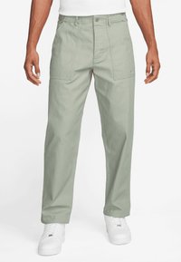 Light green trousers feature a relaxed fit, textured fabric, front pockets, and a button closure. Paired with white sneakers.