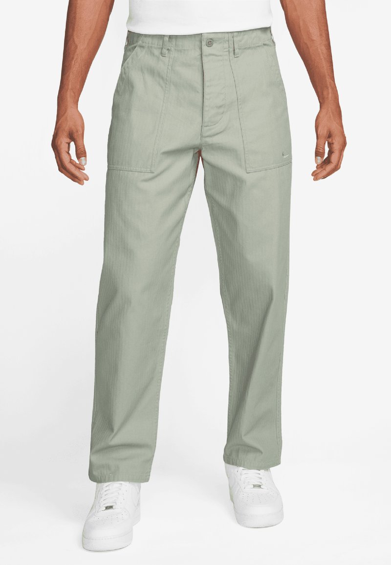 Light green trousers feature a relaxed fit, textured fabric, front pockets, and a button closure. Paired with white sneakers.