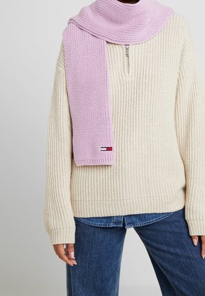 Personne portant un pull crème à côtes avec demi-fermeture éclair, un jean bleu et une écharpe tricotée lavande drapée sur l'épaule avec un petit écusson logo.