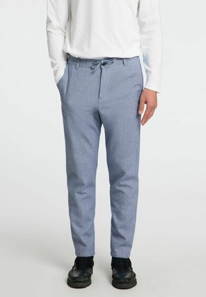 Homme portant un pantalon sur mesure bleu clair, des chaussures noires et une chemise blanche à manches longues, debout avec une main dans la poche.