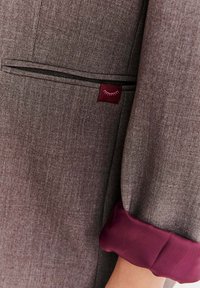 Szary marynarka o teksturowanej powierzchni z bordowymi mankietami i detalem kieszeni z małym logo. Zbliżenie podkreślające tkaninę i wykonanie.