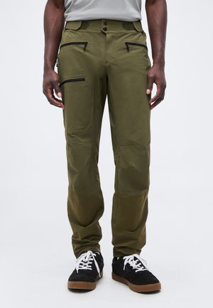 Vyras vilki alyvuogių žalumos krovinių kelnes su juodais užtrauktukų kišenėmis ir juodus sportinius batelius su baltais raišteliais, stovintis prieš paprastą foną.