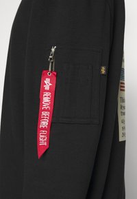 Manche noire avec une poche rectangulaire zippée et une étiquette en tissu rouge portant la mention « Remove Before Flight » attachée au curseur de la fermeture éclair.