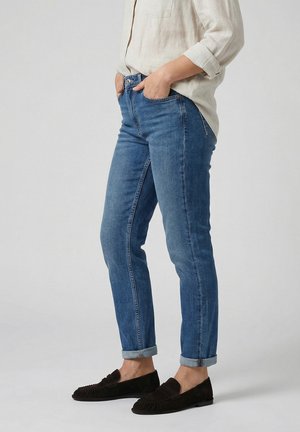 Osoba nosící modré džíny s obrubou, béžové tričko s dlouhým rukávem a srolovanými rukávy a tmavě hnědé nazouvací boty, stojící s rukama v kapse.