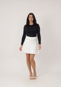 Mujer con cabello largo y oscuro, vestida con top negro de manga larga, falda plisada blanca y tacones abiertos blancos, de pie contra un fondo liso.