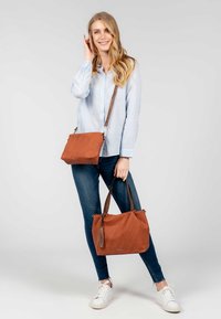 Femme en chemise rayée bleue et jeans tenant deux sacs à main marron, un porté en bandoulière et un fourre-tout, souriant devant un fond clair uni.