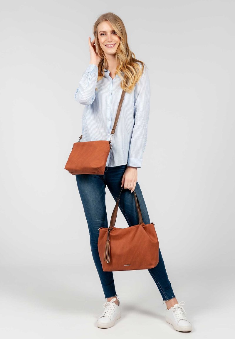 Femme en chemise rayée bleue et jeans tenant deux sacs à main marron, un porté en bandoulière et un fourre-tout, souriant devant un fond clair uni.