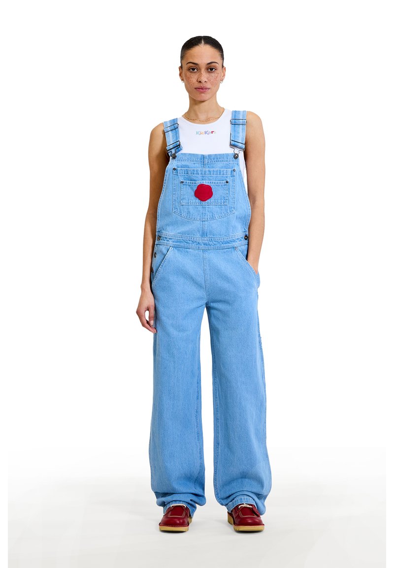 Overoles de mezclilla azul claro con piernas anchas, tirantes ajustables, bolsillo delantero y un parche circular rojo en el pecho. Combinados con zapatos rojos.