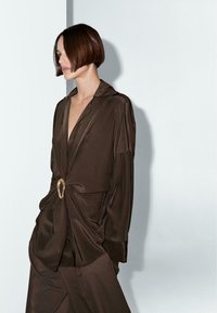 Tenue en satin marron composée d'un haut ajusté avec un décolleté plongeant, des manches longues et un accent décoratif en métal doré à la taille.