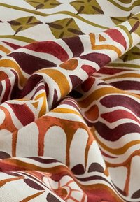 Tissu présentant un motif coloré de formes organiques en rouge, orange, vert et marron sur un fond crème. Le matériau semble texturé.