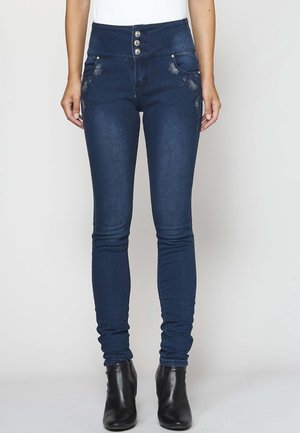 Frau trägt dunkelblaue High-Waist-Skinny-Jeans mit Blumenstickerei auf den Taschen und schwarze Knöchelstiefel, steht vor weißem Hintergrund.