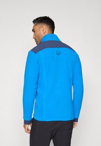 Giacca in fleece di un blu intenso con una superficie testurizzata, dettagli in blu navy sulle spalle, colletto alto e logo sul retro.