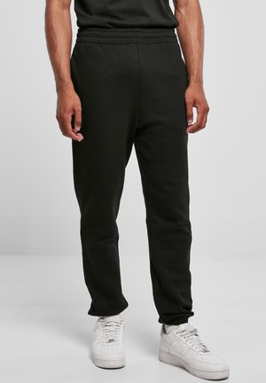 Man met zwarte joggerbroeken met elastische taille en witte sneakers, staand tegen een neutrale achtergrond.