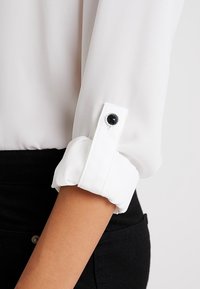 Camicia bianca con maniche arrotolate, caratterizzata da un bottone decorativo nero e un tessuto morbido e leggero. Pantaloni neri visibili nella parte inferiore.