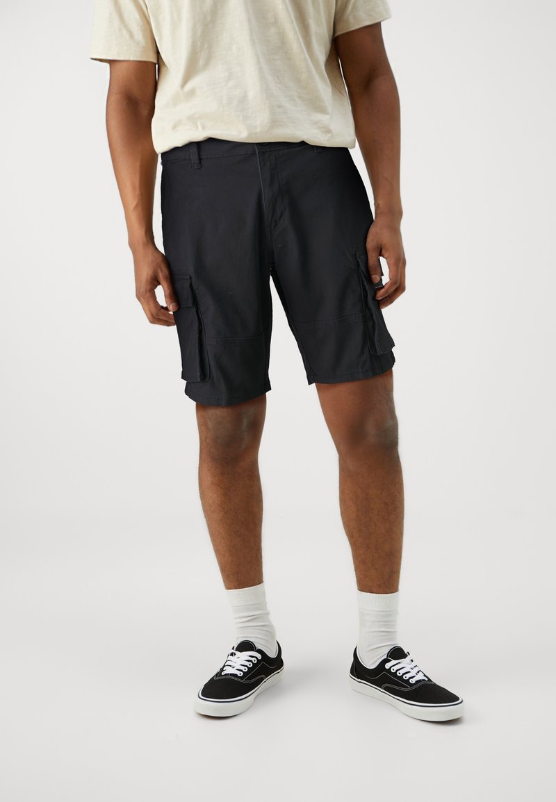 Shorts cargo noirs avec deux poches latérales, fabriqués en tissu léger. Portés avec un t-shirt beige clair et des baskets noires avec des accents blancs.