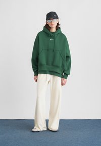 Person som bærer en mørkegrønn Nike hettegenser, hvite løse bukser, grå sneakers, grå solbriller og en svart Nike caps mens de står på et blått teppe.