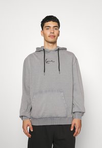 Grauer Kapuzenpullover aus weichem Stoff. Verfügt über eine Vordertasche, elastische Bündchen und ein schwarzes, gesticktes Logo auf der Brust.