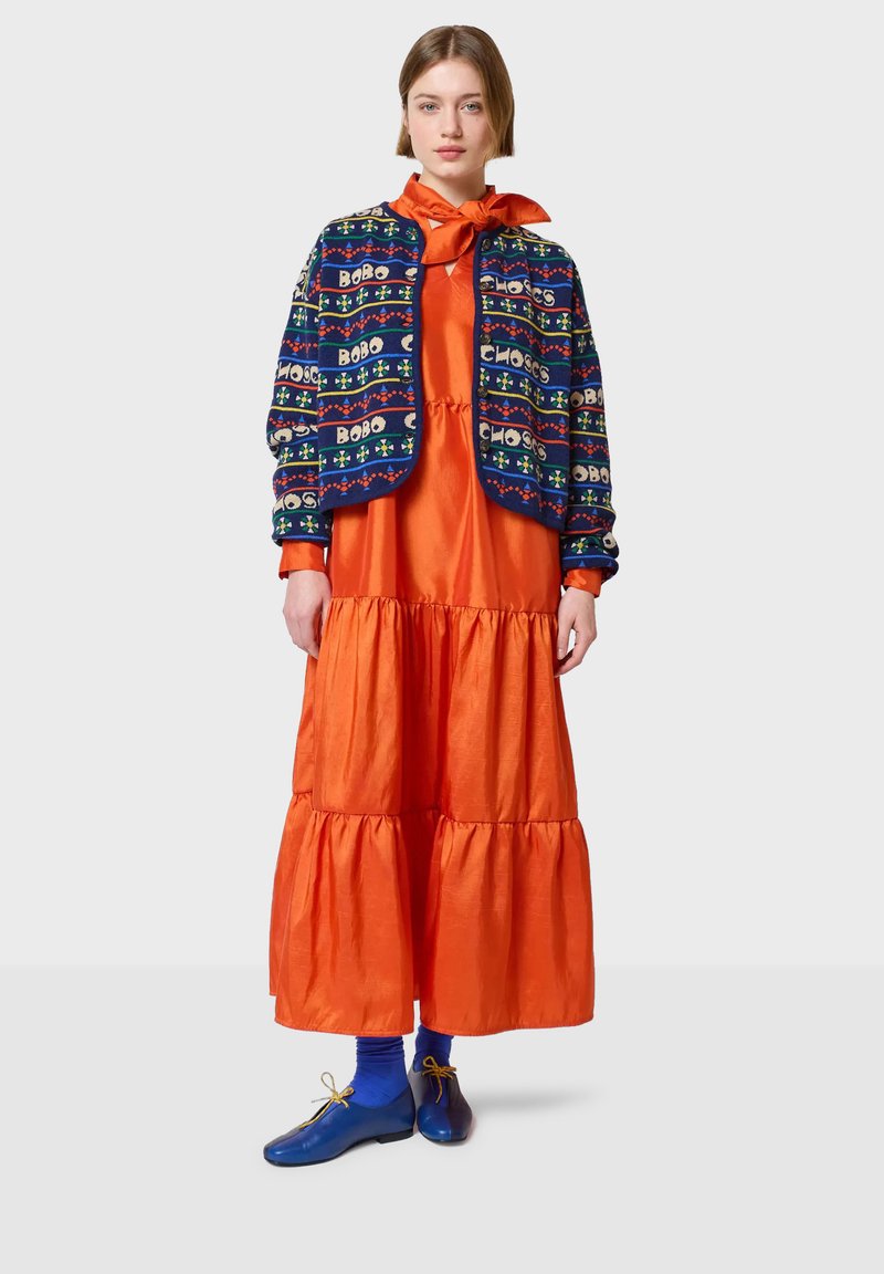 Chaqueta recortada colorida con patrones de texto audaces, sobre un vestido maxi naranja de volantes. Zapatos azules con cordones completan el conjunto.