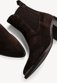 Bottines en daim marron foncé avec bouts pointus, panneaux latéraux élastiques et petits talons blocs sur fond blanc.