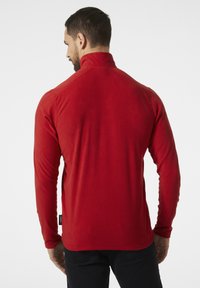 Czerwony polarowy pulower z długim rękawem i wysokim kołnierzem. Posiada szwy płaskie oraz subtelną metkę marki na dolnej stronie. Gładka tekstura.