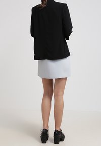 Kobieta w czarnym żakiecie, białej mini spódnicy i czarnych butach na platformie, stojąca z plecami zwrócona w stronę aparatu na jednolitym tle.