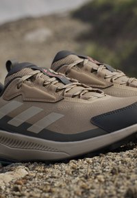 Buty do biegania w terenie w kolorze brązowym z siateczki z ciemnoszarymi akcentami. Wśród cech produktu znajdują się teksturowana cholewka, wzmocniony czubek buta oraz wzorzyste sznurowadła.
