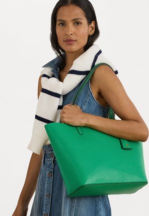 Zöld bőr tote táska texturált felülettel, egyszerű dizájnnal és masszív fogantyúkkal. A modellen farmerruha és csíkos pulóver van.