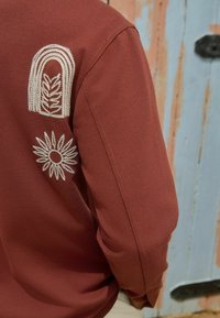 Marlot-langærmet skjorte med tekstureret stof. Har hvid broderi af en bue og et blomstermønster på venstre arm.