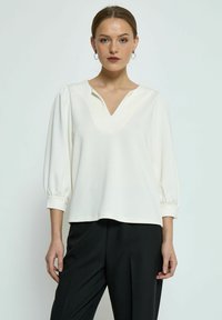 Witte pullover met een V-hals, driekwart pofmouwen en een gestructureerde stof. Gecombineerd met zwarte trousers, tegen een neutrale achtergrond.