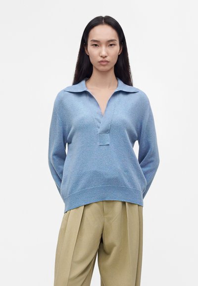 Femme aux longs cheveux foncés porte un pull bleu clair à col et un pantalon beige plissé, debout devant un fond blanc uni.