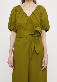 Olivově zelené šaty s výstřihem do V a nafouknutými rukávy. Mají opasek na zavázání v pase a hladkou, strukturovanou tkaninu. Jednoduchá a elegantní silueta.