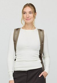 Bruine rugzak met gevoerde, verstelbare schouderbanden. Model draagt een aansluitend wit langemouwen shirt en donkere broek, glimlachend.