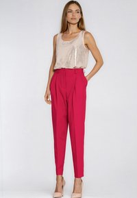 Bezrekawnik z cekinów w jasnoróżowym kolorze zestawiony z wysokim stanem jasnofuksjowych plisowanych spodni. Modelka ma na sobie beżowe szpilki.