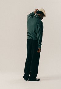 Άνδρας φοράει μπεζ καπέλο, σκούρο πράσινο πουλόβερ, μαύρο φαρδύ παντελόνι, χρυσό σκουλαρίκι κρίκος, γυαλιά και παπούτσια, στέκεται μπροστά σε απλό φωτεινό φόντο.