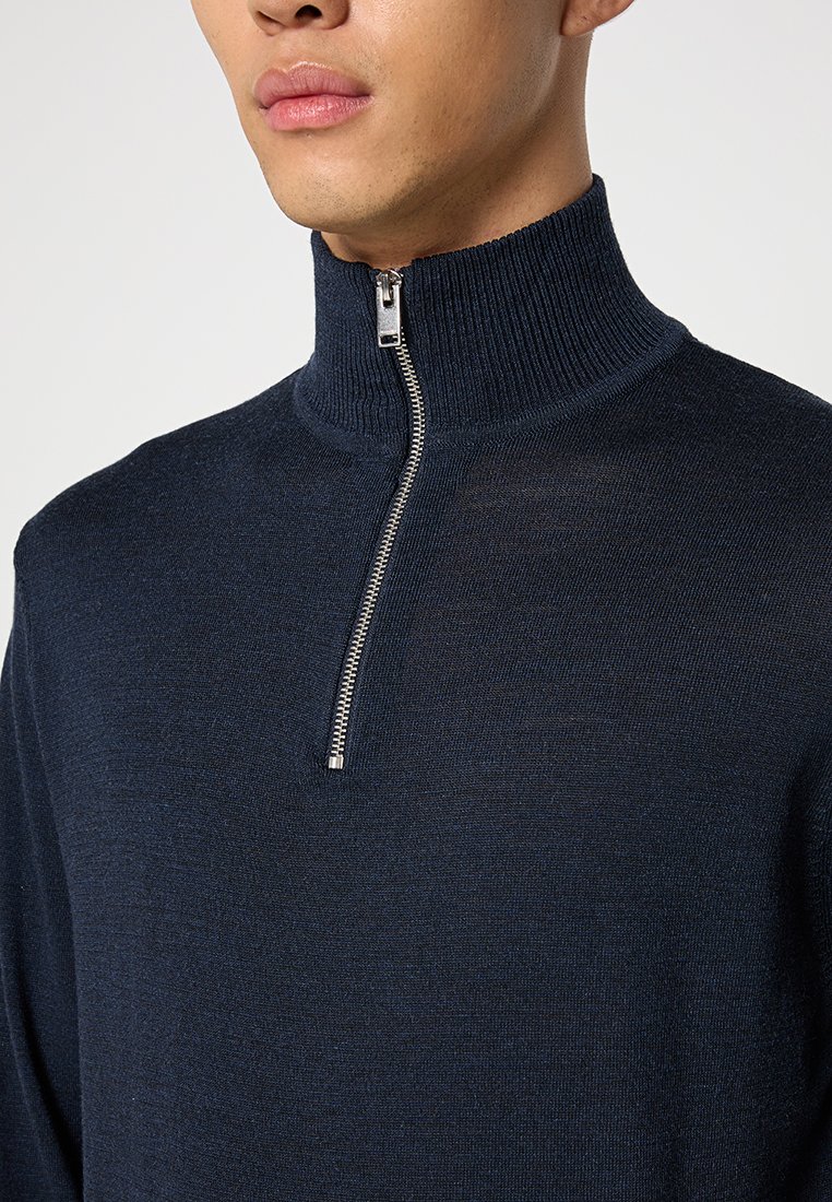 Marine gebreide pullover met hoge kraag, voorzien van een zilveren ritsluiting en geribbelde afwerking bij de hals en mouwen.