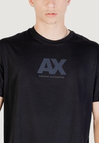 T-shirt en coton noir avec un grand logo "AX" gris et le texte "ARMANI EXCHANGE" dans un style contrasté sur le devant. Coupe régulière.