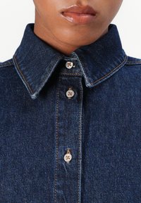 Dark blue denim shirt with a classic collar, visible stitching, and metallic buttons. The fabric has a smooth texture and a structured shape.
