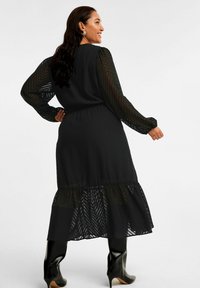 Schwarzes Kleid mit langen transparenten Ärmeln, strukturiertem Stoff, gestuftem Saum, elastischem Bund, kombiniert mit glänzenden knielangen Stiefeln.