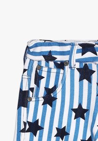 Białe dżinsy z niebieskimi paskami i czarnymi wzorami gwiazd. Wyposażone w sprzęt w kolorze srebrnym oraz standardowy krój pięciu kieszeni.