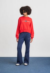 Červený skrátený sweatshirt s logom "Nike Shox", kombinovaný s tmavomodrými širokými nohavicami s bielymi pruhmi po stranách. Čierne slnečné okuliare, minimalistická obuv.