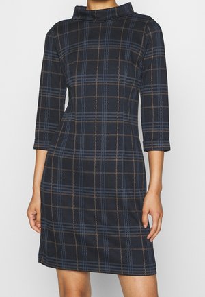 Femme portant une robe à carreaux bleu foncé et noir avec des manches trois-quarts et un col montant, debout les bras relâchés le long du corps.
