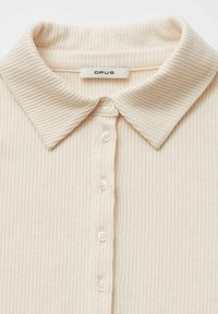 Polo beige a coste con colletto classico, abbottonatura frontale con cinque bottoni e tessuto morbido. Marchio "OPUS".