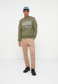 Felpa verde oliva con testo grafico bianco e blu, abbinata a pantaloni beige chiaro e sneakers nere, e un cappellino di jeans.
